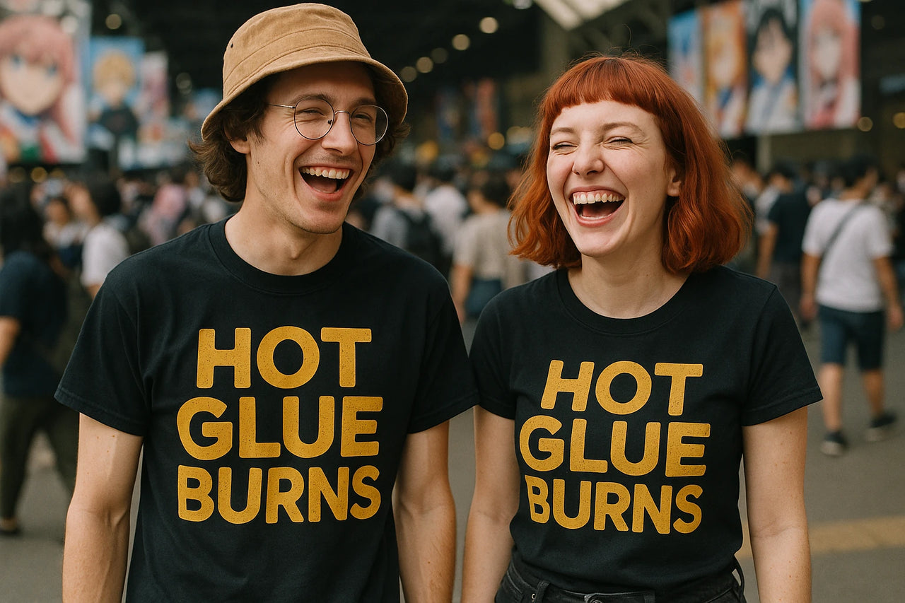Two people are standing side by side, both laughing and wearing matching black t-shirts with the text "HOT GLUE BURNS" in bold yellow letters. The person on the left is wearing glasses and a bucket hat, while the person on the right has short, red hair. They appear to be in a crowded indoor setting.