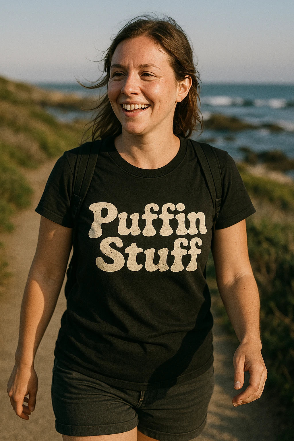 A smiling person wearing a black T-shirt with white text that says "Puffin Stuff." They are outdoors on a path near the coastline, with the ocean and greenery visible in the background. The person has long brown hair and is wearing a backpack.