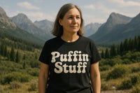 A person stands outdoors in front of a mountainous landscape wearing a black t-shirt with the text 