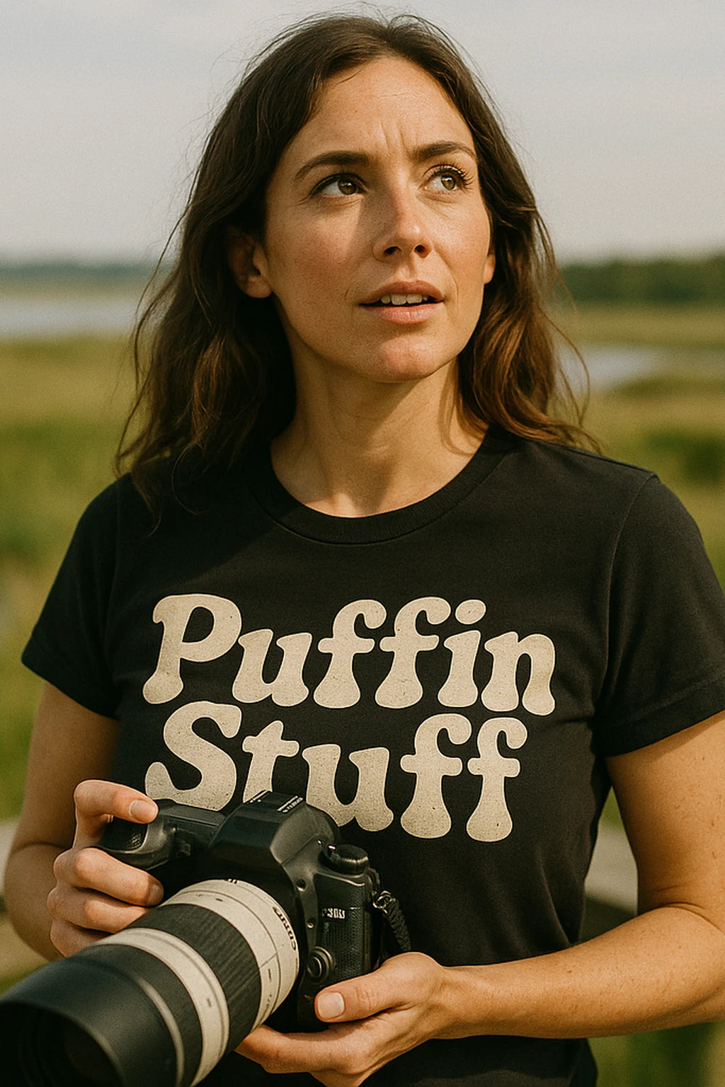 A person with long brown hair is holding a camera and looking off into the distance. They are wearing a black T-shirt with the text 'Puffin Stuff' on it. The background features a natural, grassy outdoor setting.