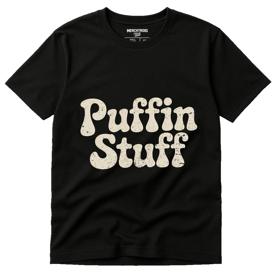 A black t-shirt featuring the words "Puffin Stuff" in large, bold, cream-colored letters across the front. The text has a slightly distressed look, giving it a vintage style. The shirt label at the neck reads "Merchtroid."