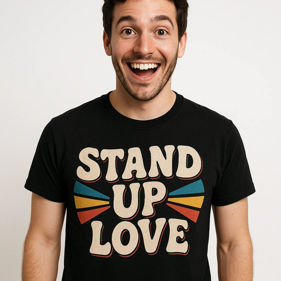 A person with a beard and dark hair is wearing a black t-shirt with the text "STAND UP LOVE" in bold, retro-style font. The person is smiling widely, with a cheerful expression. The design on the shirt includes colorful shapes on either side of the text.