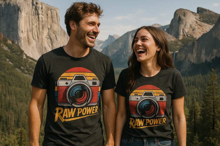 A man and woman are standing outdoors, wearing matching black t-shirts with a retro camera design and the text "Raw Power." They are smiling and appear to be in a mountainous setting with trees and rock formations in the background.