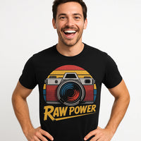 A person with short dark hair and a beard is smiling widely, wearing a black t-shirt featuring a retro-style graphic of a camera with a colorful background and the text 