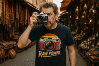 A person with short hair and a beard is holding a camera up to their eye in a bustling market setting with rugs and brassware. They are wearing a black t-shirt featuring an illustration of a camera and the text 
