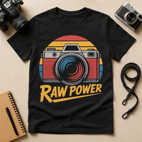 A black t-shirt featuring a graphic of a vintage camera with a retro color scheme of red, yellow, and blue. Surrounding items include two cameras, a camera strap, and a notebook with a pen.
