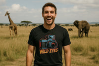 A man is standing in a grassy savanna, smiling widely. He is wearing a black t-shirt with an illustration of a camera and the words 