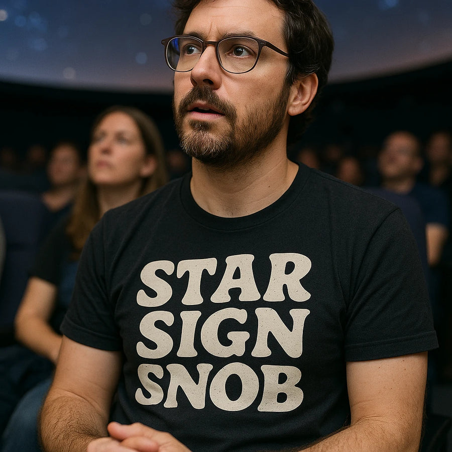 A person with a beard and glasses is wearing a black T-shirt with the text "STAR SIGN SNOB" printed on it. They are seated indoors with other people visible in the background, and they appear to be focused on something ahead.