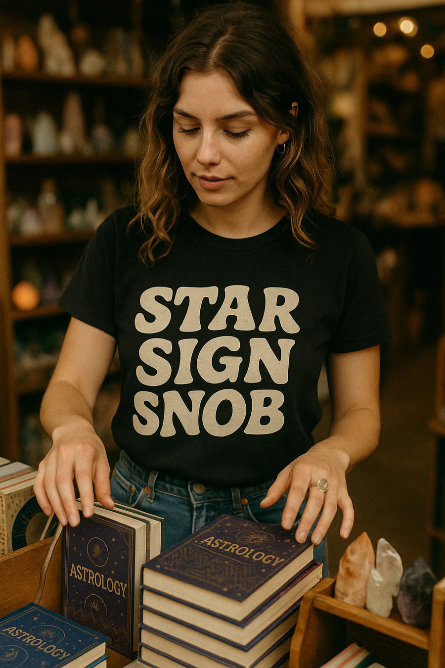 A person with wavy brown hair is wearing a black T-shirt that reads "STAR SIGN SNOB." They are standing in a store, arranging books titled "ASTROLOGY" on a shelf. The background features shelves with various colorful items, possibly crystals or decorative stones.