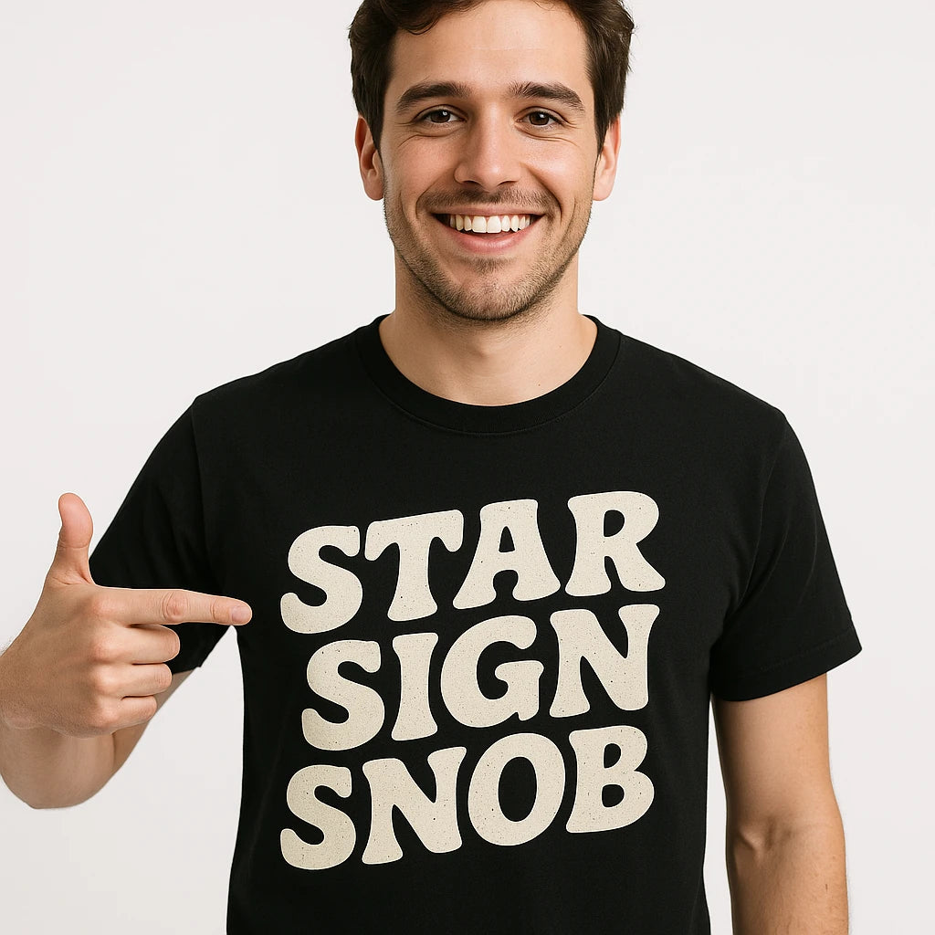A person smiling and pointing at the text on their black T-shirt which reads "STAR SIGN SNOB." The individual has short dark hair and is standing against a plain background.