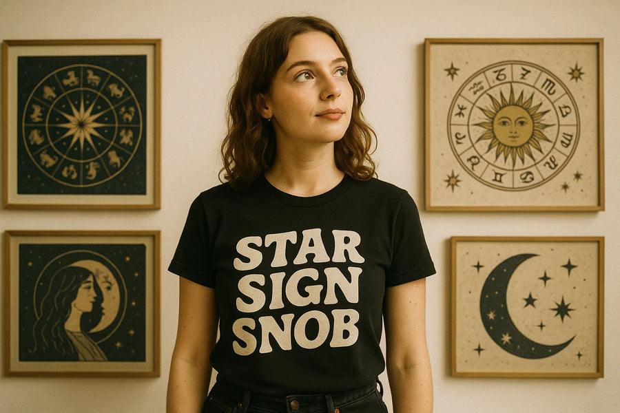 A person with shoulder-length brown hair is wearing a black t-shirt with the words "STAR SIGN SNOB" printed in bold, white letters. Behind them, there are framed artworks depicting zodiac and celestial themes, including designs of a zodiac wheel, a sun with a face, a crescent moon with stars, and a woman's profile combined with a crescent moon.