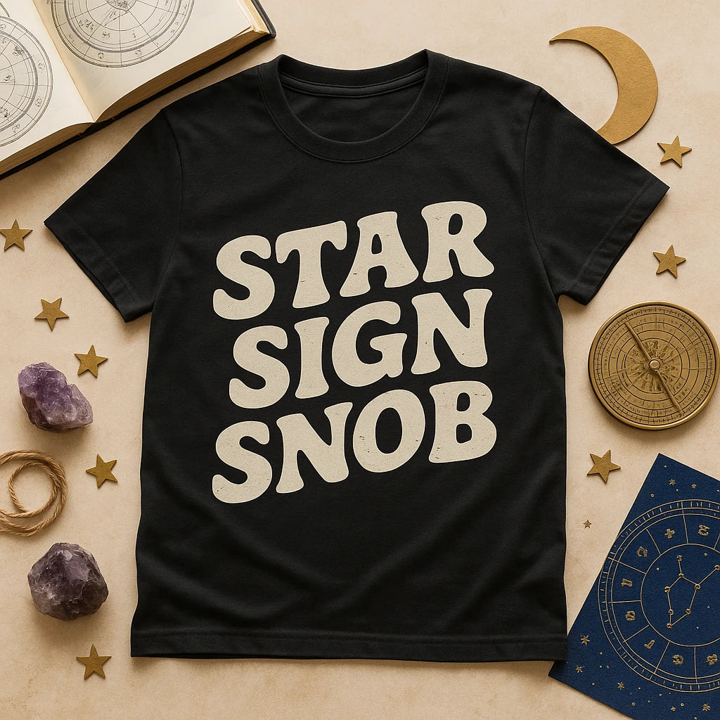A black T-shirt with bold white text that reads "STAR SIGN SNOB" is laid out on a table. Around the T-shirt, there are various astrological and celestial themed items, including an open book with star charts, golden stars, amethyst crystals, a rope, a golden crescent moon, a blue card with a zodiac wheel, and a brass compass-like object.