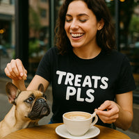 A person with wavy brown hair is sitting at a table with a cup of coffee, smiling and holding one ear of a small, light brown dog with pointed ears. The person is wearing a black t-shirt with the text 