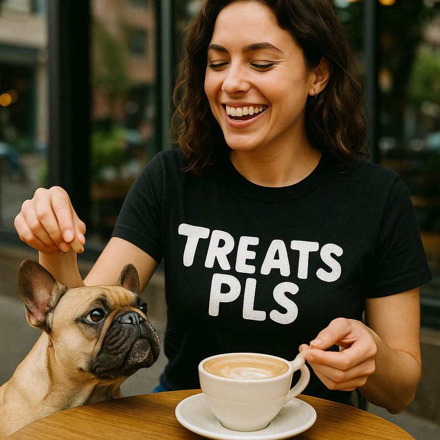 A person with wavy brown hair is sitting at a table with a cup of coffee, smiling and holding one ear of a small, light brown dog with pointed ears. The person is wearing a black t-shirt with the text "TREATS PLS" printed on it. The setting appears to be outdoors at a cafe.