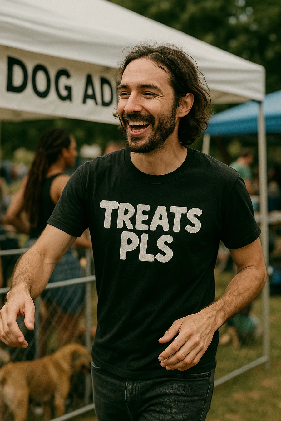 A smiling person with long hair and a beard is wearing a black t-shirt with the text "TREATS PLS." They are standing outside near a white tent with the partial text "DOG AD" visible, likely at an outdoor event related to dogs. There are other people and a dog in the background.