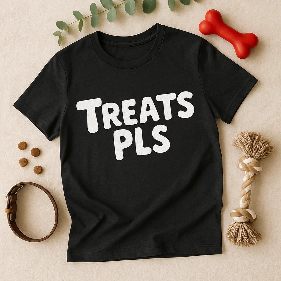 A black T-shirt with the text "TREATS PLS" in bold white letters is laid flat on a beige surface. Surrounding the shirt are dog-related items: a red bone-shaped toy, a rope toy, several kibble pieces, a brown leather collar, and a sprig of green leaves.