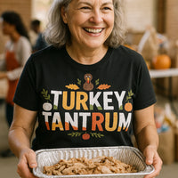 A person with gray hair is smiling while holding a tray of sliced turkey. They are wearing a black T-shirt with colorful text that reads “TURKEY TANTRUM,” and features decorative elements like a cartoon turkey and autumn leaves. The background shows a blurred setting with people and pumpkins, suggesting a festive or holiday gathering.