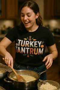 A person with long brown hair is smiling while cooking in a kitchen. They are stirring a pot on the stove. The person is wearing a black shirt with the text 