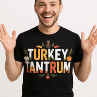 A person is smiling widely with both hands raised, wearing a black t-shirt. The shirt features colorful text that reads 