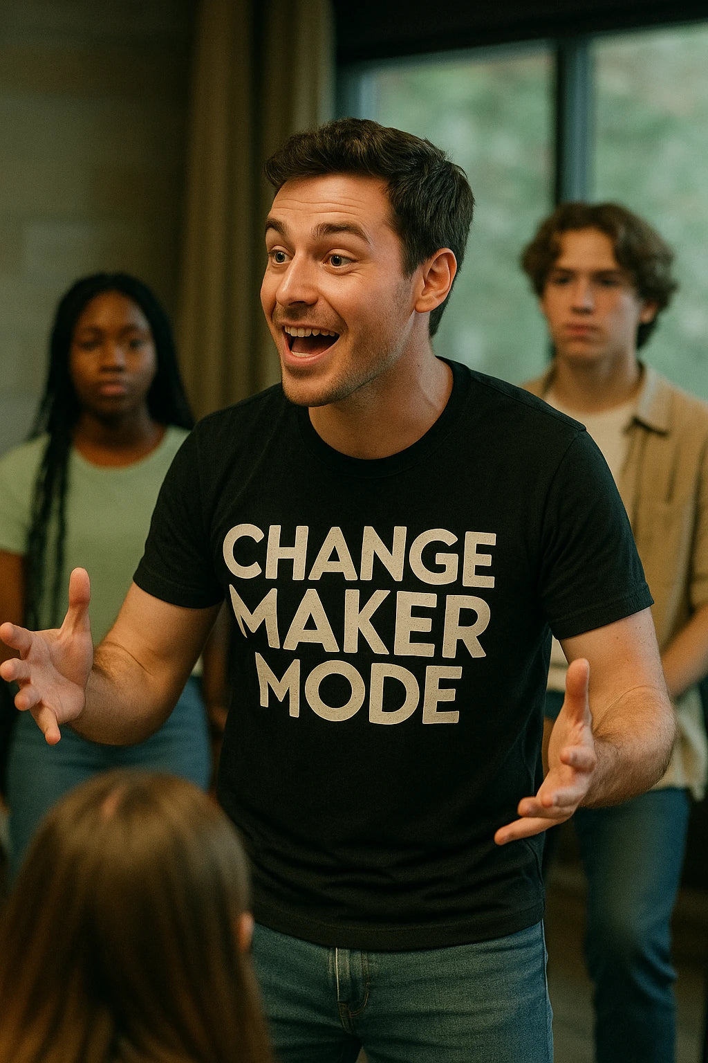 A person with short brown hair is animatedly speaking while wearing a black t-shirt with the text "CHANGE MAKER MODE." In the background, there are a few other people, including a person with long braided hair and another with medium-length wavy hair, all observing attentively.