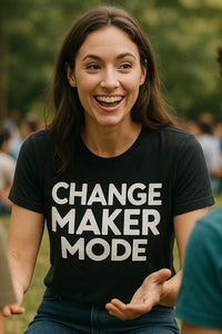 A woman with long brown hair smiles widely while sitting outdoors. She is wearing a black t-shirt with white text that reads 