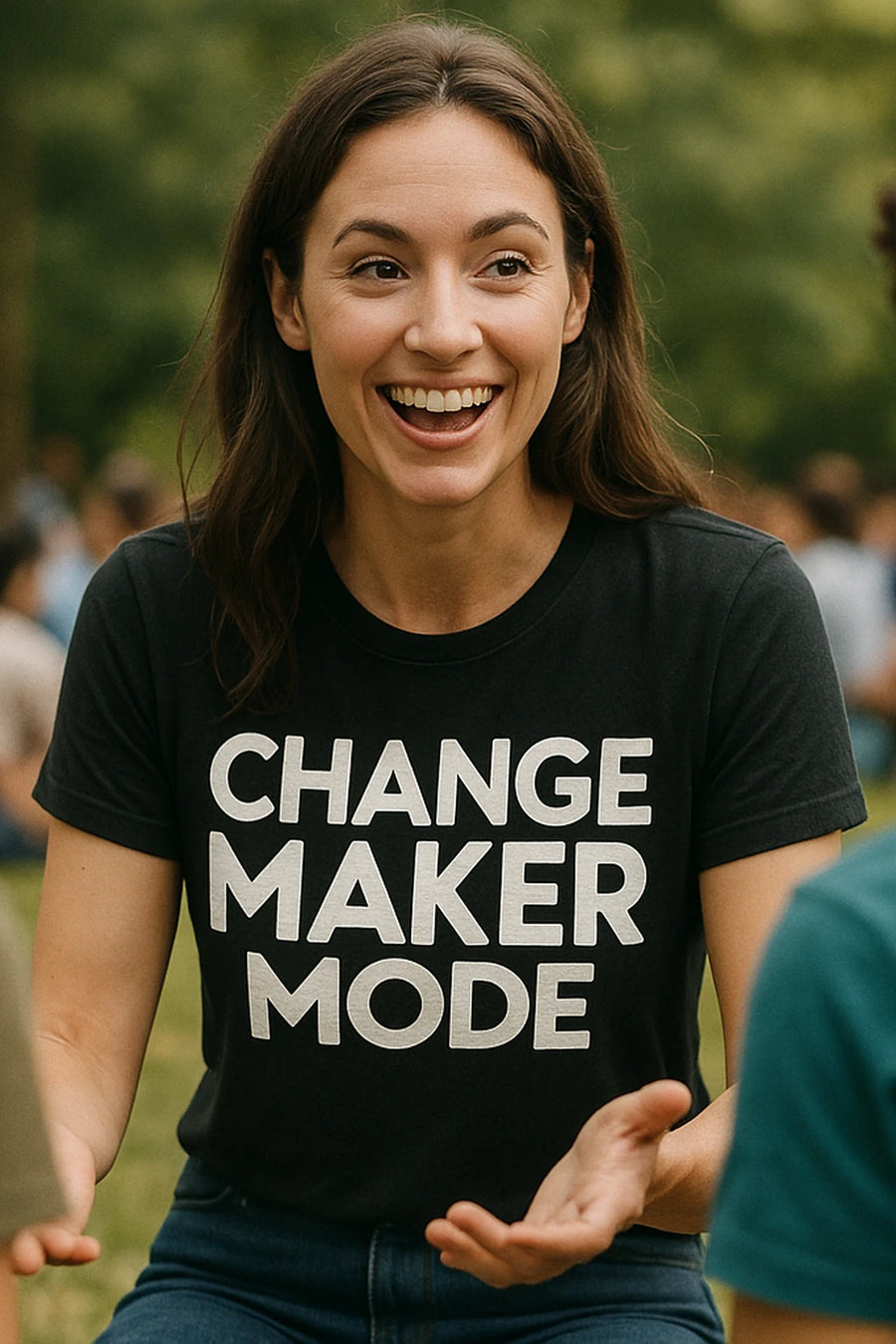 A woman with long brown hair smiles widely while sitting outdoors. She is wearing a black t-shirt with white text that reads "CHANGE MAKER MODE." She appears engaged in conversation. The background is a blurred outdoor setting with trees and people.
