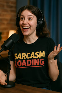 A person with long brown hair is sitting at a table with a microphone, wearing headphones and a black t-shirt that reads 
