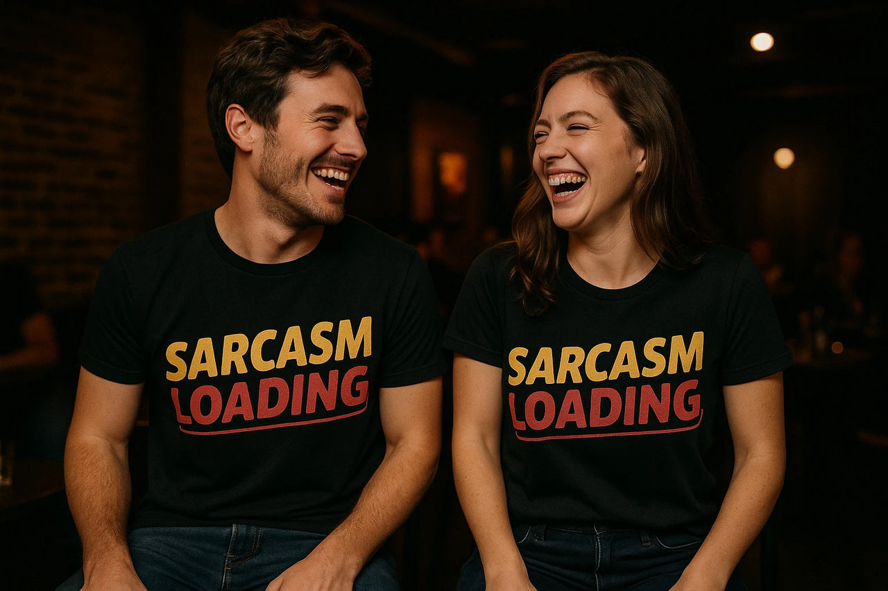 A man and a woman are sitting and laughing together. Both are wearing black T-shirts with the text "SARCASM LOADING" printed in yellow and red. They are in a dimly lit room, likely a cafe or bar.