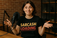 A person with short brown hair is sitting at a table with a microphone. They have a humorous expression with raised eyebrows and a slight smile, wearing a black t-shirt with the text 