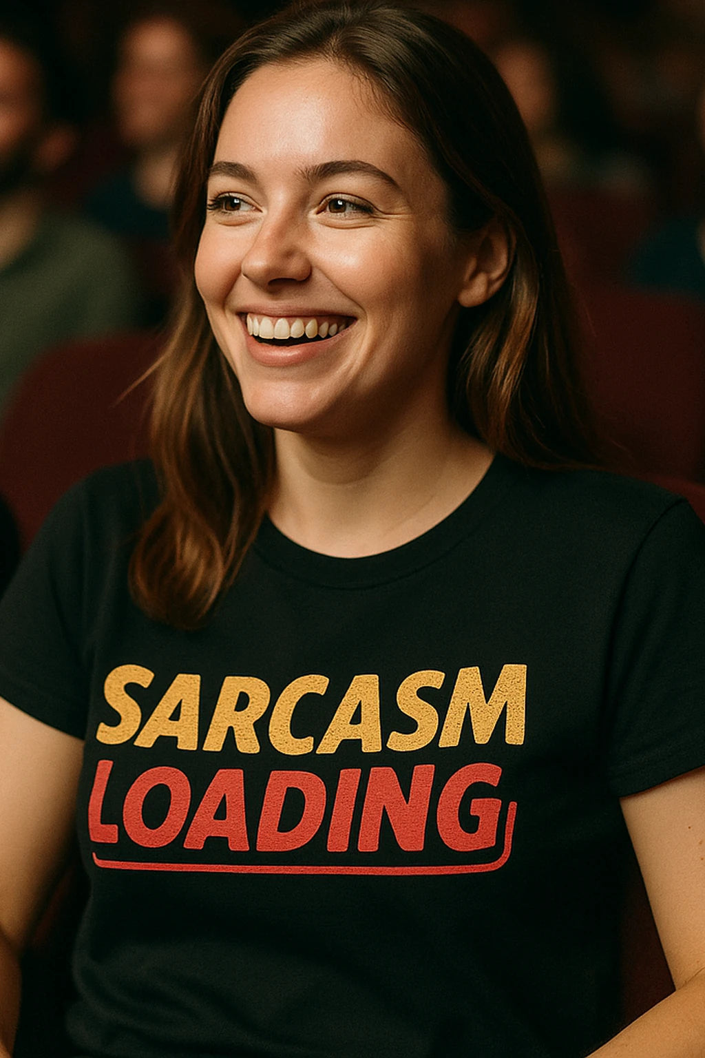 A person with long brown hair is smiling while wearing a black T-shirt with the text "SARCASM LOADING" printed on it in yellow and red letters. They are seated indoors with other people in the background.