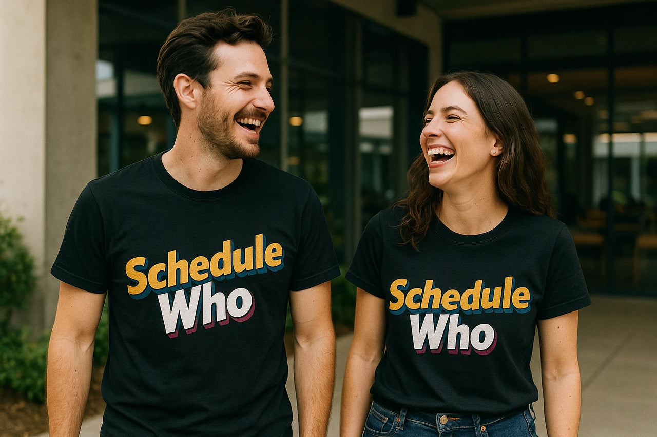 A man and a woman are standing side by side, both laughing and wearing matching black T-shirts with the text "Schedule Who" printed on them. They are outdoors, with a building featuring large windows in the background. The man has short dark hair and a beard, while the woman has long wavy hair.