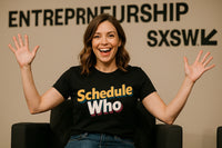 A person with shoulder-length brown hair is sitting in a chair, smiling broadly with their hands raised. They are wearing a black T-shirt with the text 