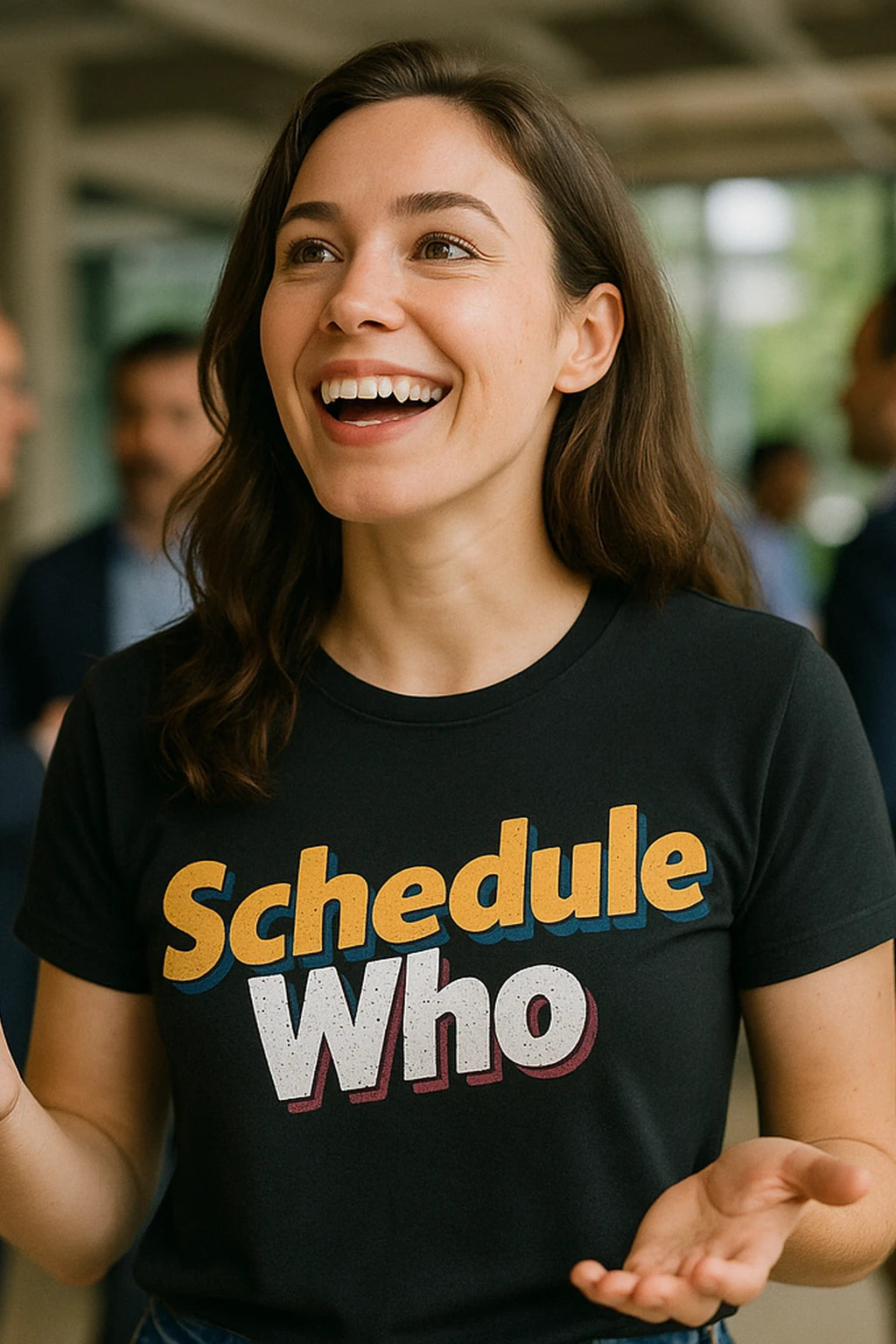 A person with long brown hair is smiling widely and wearing a black t-shirt with the text "Schedule Who" printed in colorful letters. The background shows a blurred indoor environment with people in the distance.
