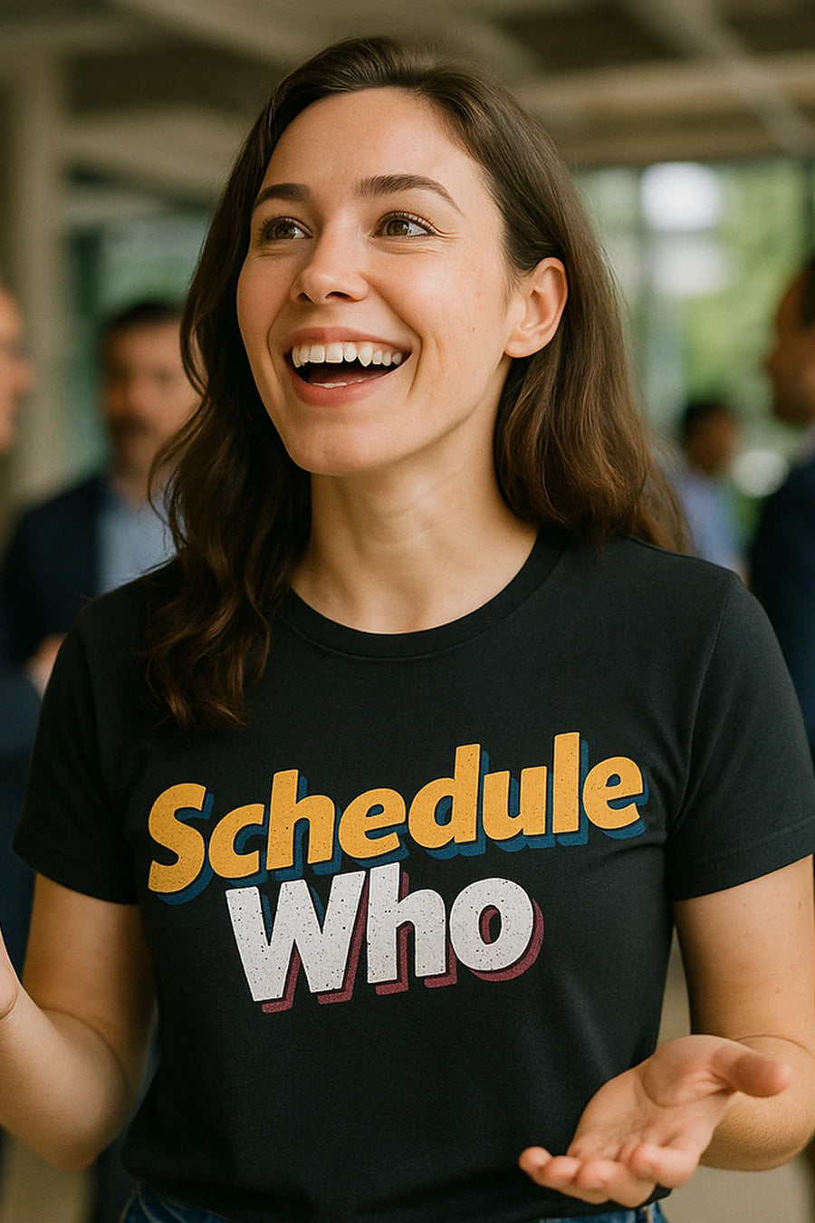 A person with long brown hair is smiling widely and wearing a black t-shirt with the text "Schedule Who" printed in colorful letters. The background shows a blurred indoor environment with people in the distance.
