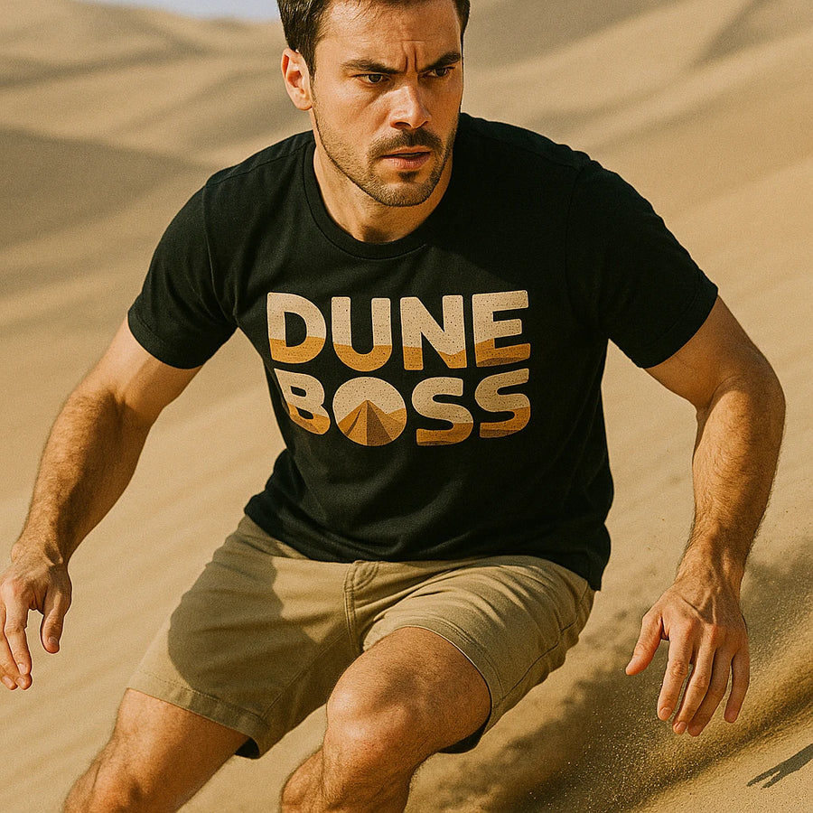 A person with short dark hair is running on sand dunes, wearing a black t-shirt with the words "DUNE BOSS" printed in bold lettering, along with a stylized dune graphic. They also wear light-colored shorts. The background features sandy hills under a clear sky.