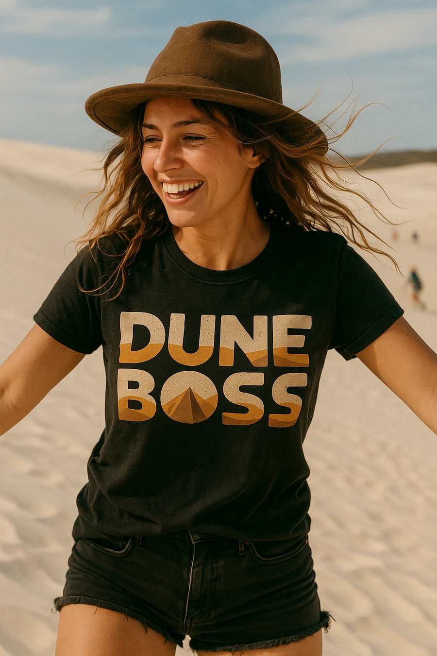 A person with long hair and a wide-brimmed hat smiling in a sandy desert environment. They are wearing a black t-shirt with the words "DUNE BOSS" printed on it and dark shorts. The sky is clear and blue.
