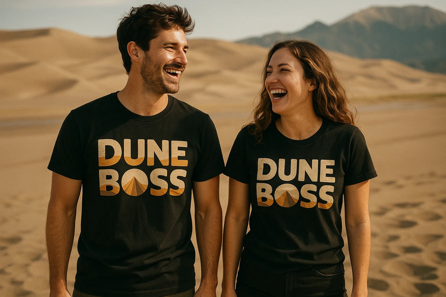 A man and a woman are standing on sand dunes, both wearing black T-shirts with the text "DUNE BOSS" in bold letters. They are looking at each other and laughing, with mountains visible in the background.