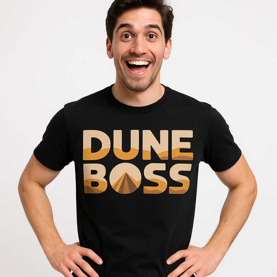 A person with short dark hair and stubble, wearing a black t-shirt with the words "DUNE BOSS" printed in bold letters. They are smiling widely and have their hands on their hips, posing against a plain white background.