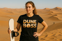 A person stands confidently on a sand dune in the desert, holding a sandboard. They have light brown hair pulled back and are wearing a black t-shirt with the text 