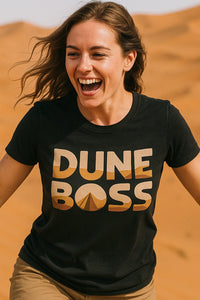 A person with long hair is smiling and running in a desert landscape, wearing a black t-shirt with 