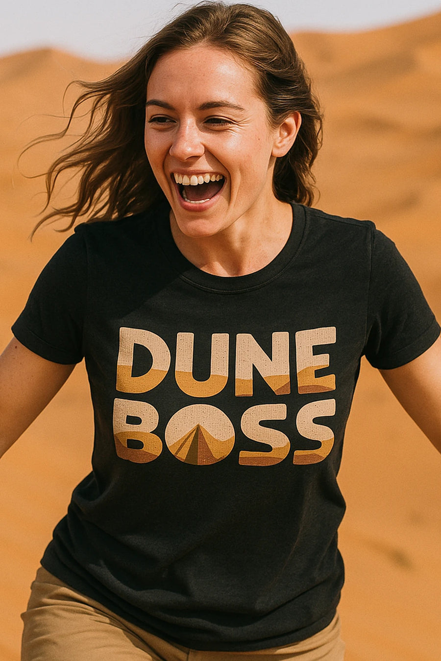 A person with long hair is smiling and running in a desert landscape, wearing a black t-shirt with "DUNE BOSS" written on it in bold letters.
