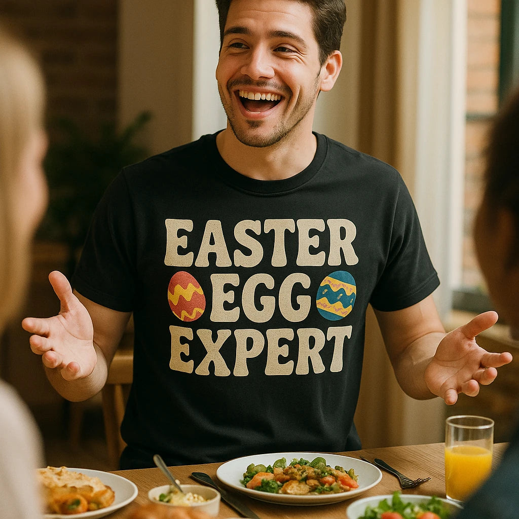 A person with short dark hair and a beard is sitting at a dining table, smiling and gesturing with their hands. They are wearing a black T-shirt with the text "EASTER EGG EXPERT" and images of colorful Easter eggs. There are plates of food and a glass of orange juice on the table.