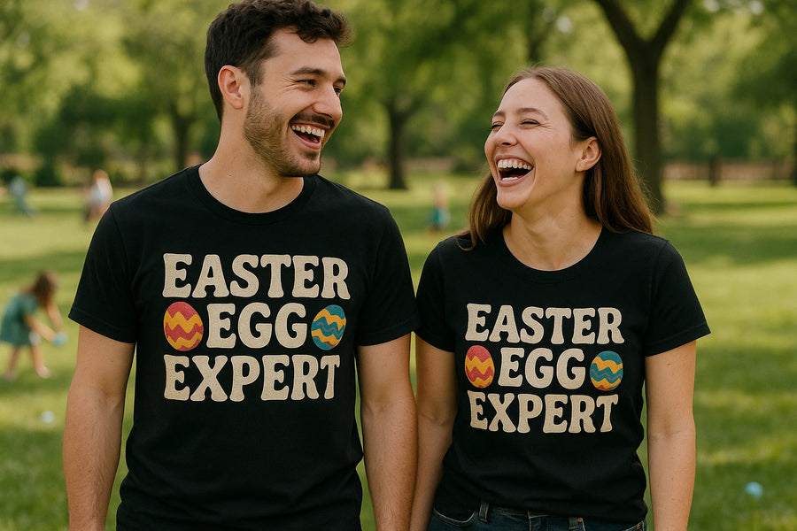 A man and woman are standing side by side in a park, both wearing black T-shirts with the text "EASTER EGG EXPERT" printed on them, accompanied by colorful images of Easter eggs. They are smiling and laughing, with a blurred background of green trees and grass.
