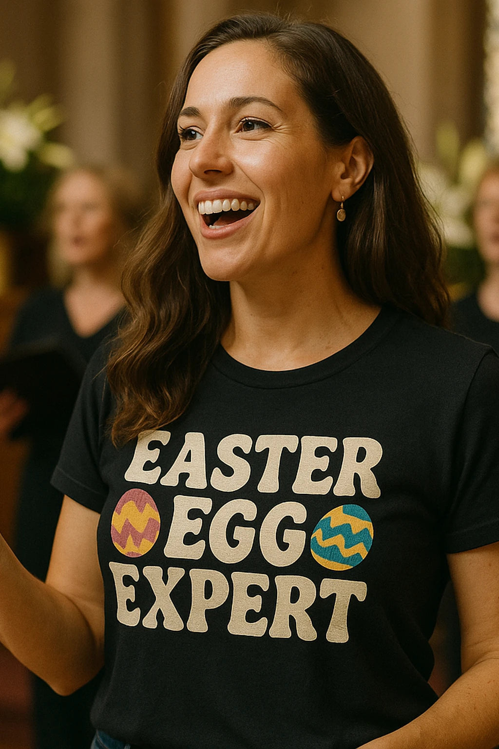 A person with long brown hair is smiling widely, wearing a black T-shirt that reads "EASTER EGG EXPERT" with colorful egg designs. They appear to be indoors, with blurred figures and floral decorations in the background.