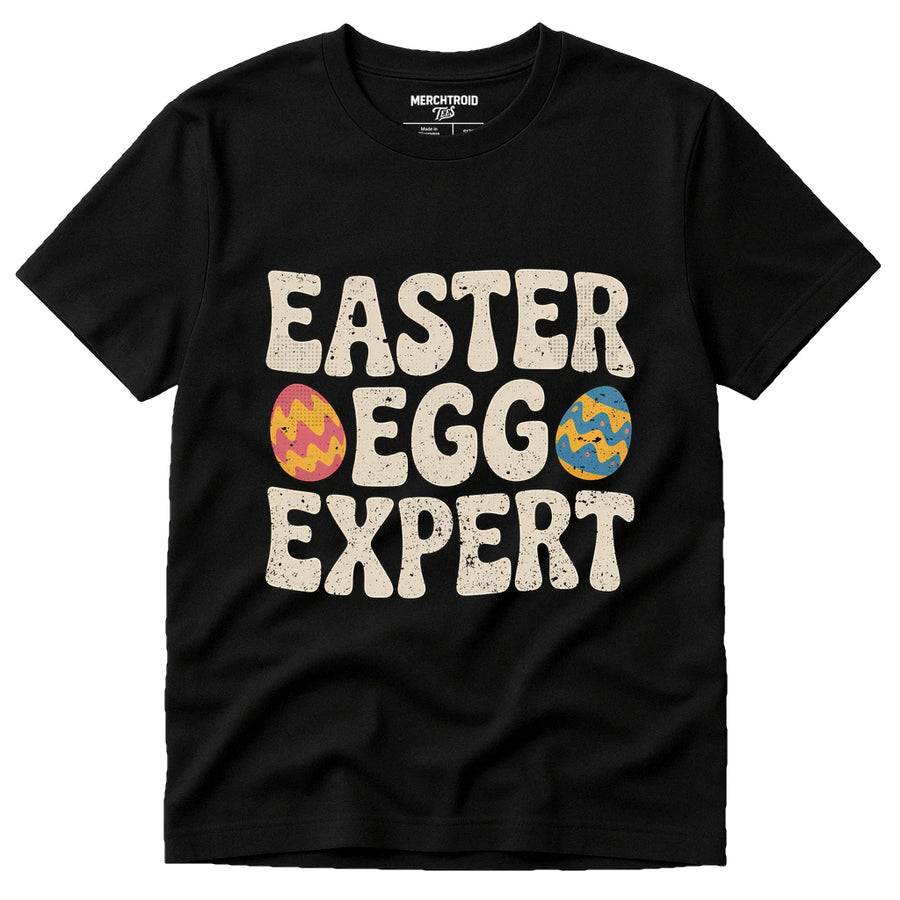 A black T-shirt featuring the slogan "EASTER EGG EXPERT" in large, bold white letters, with colorful Easter eggs integrated into the letters "E" and "G".