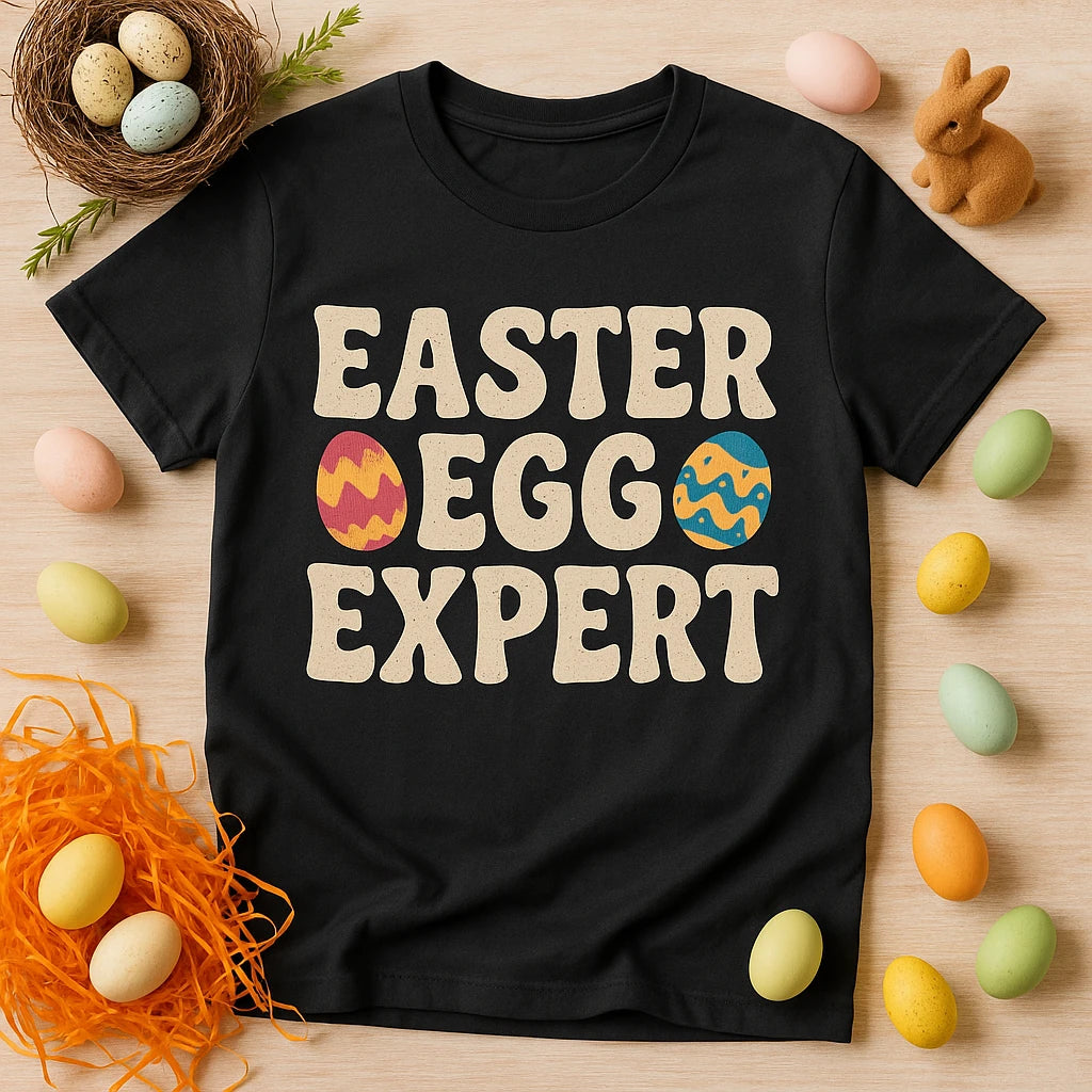 A black t-shirt laid flat, featuring the text "EASTER EGG EXPERT" with decorated egg graphics on the words "EGG". The shirt is surrounded by pastel-colored eggs, a small nest with three eggs, orange shredded paper, and a small brown rabbit figurine placed on a wooden surface.