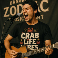 A person wearing a backward cap passionately sings into a microphone while playing an acoustic guitar. They are wearing a black T-shirt with a crab design and the text 