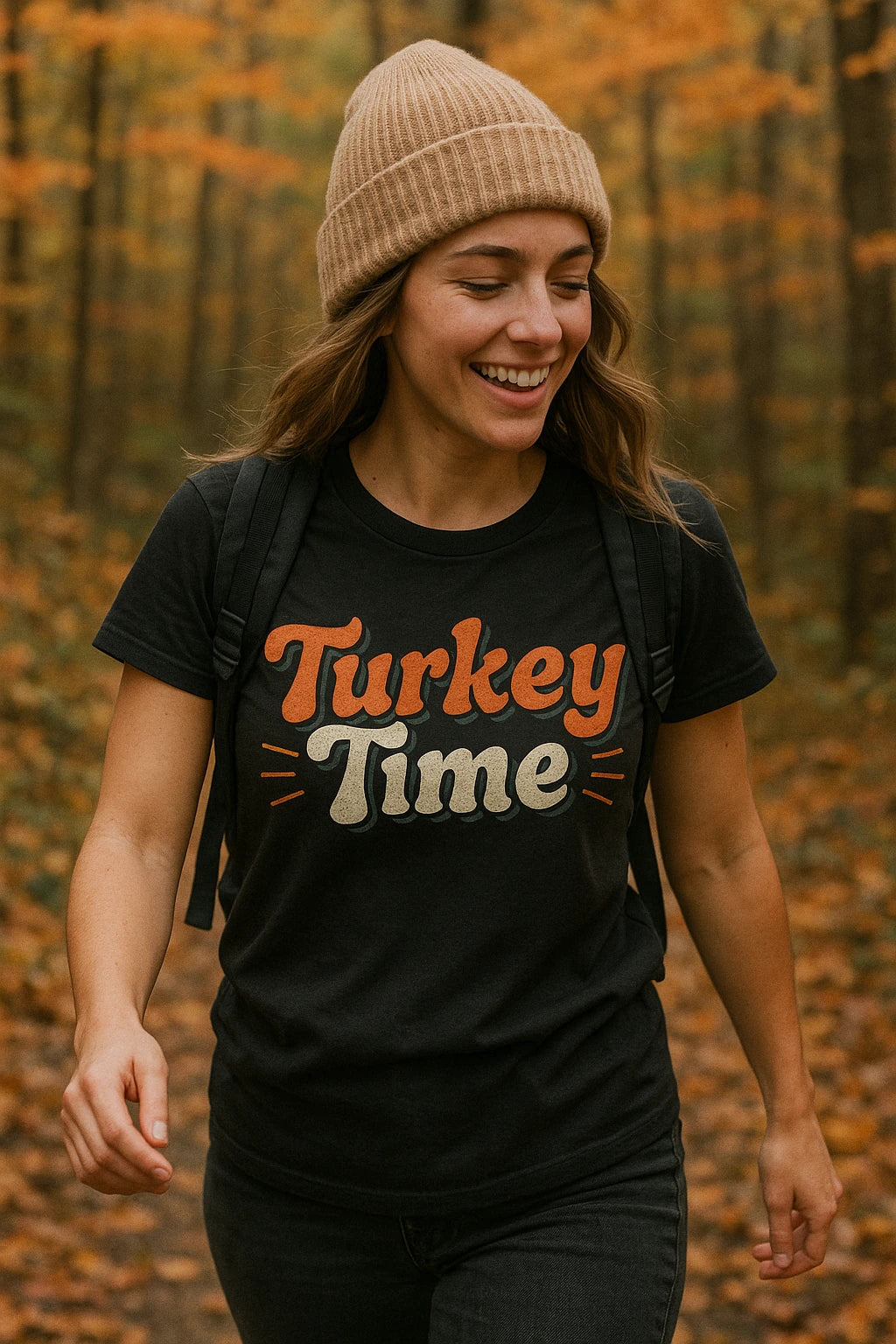 A person wearing a black t-shirt with "Turkey Time" written on it, a beige knit hat, and a backpack. They are walking through a forest with orange and yellow autumn leaves. The person is smiling and appears to be enjoying the scenic outdoors.
