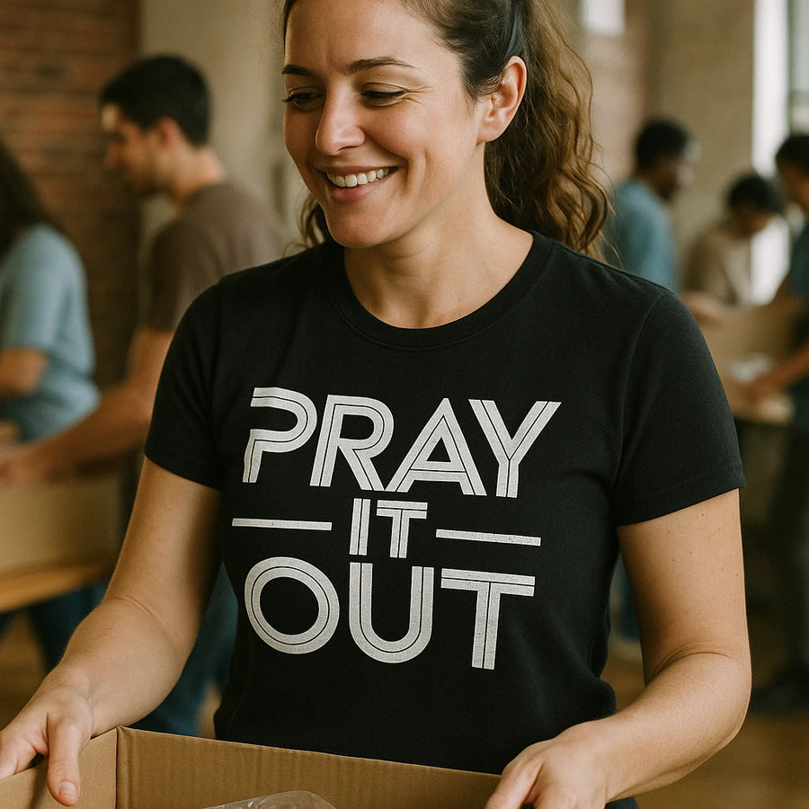 A person with long hair in a ponytail is wearing a black t-shirt that reads "PRAY IT OUT." They are smiling and holding a cardboard box in a room where others are engaged in similar activities.