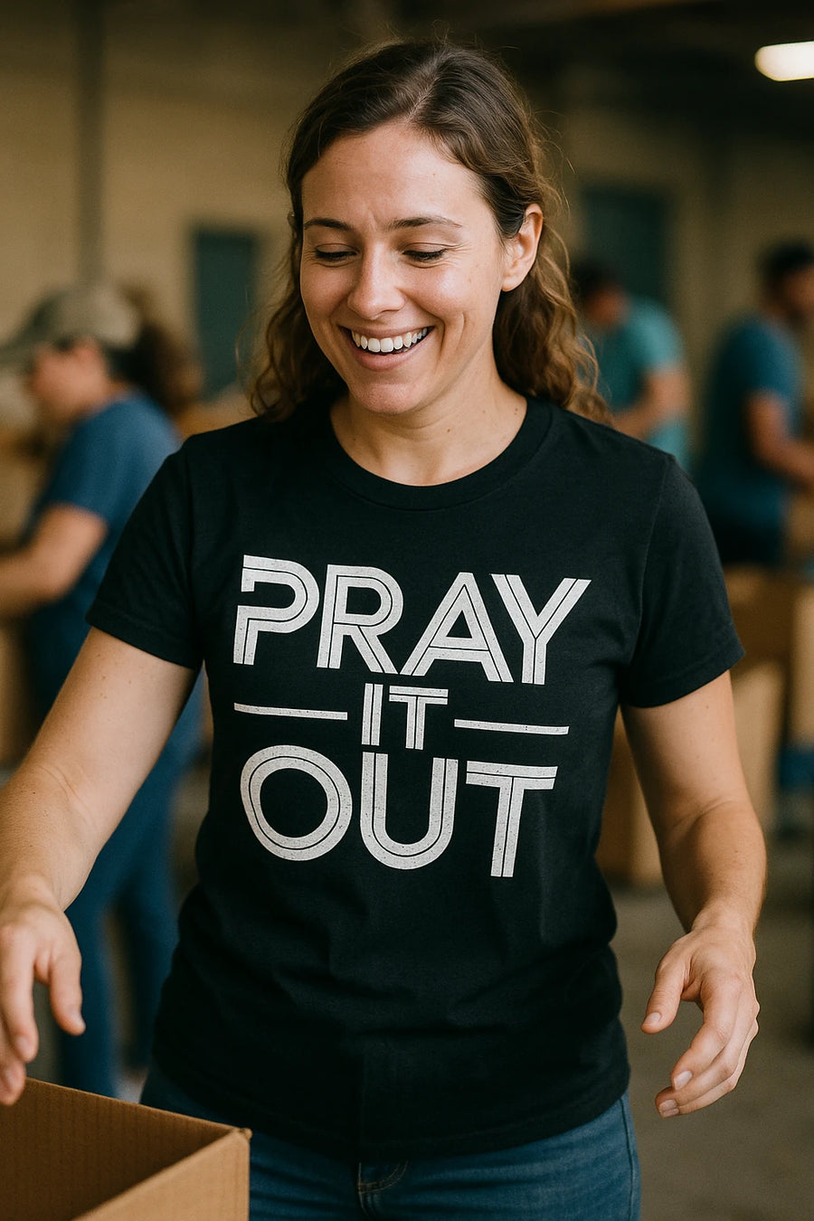 A person with long brown hair is smiling and wearing a black t-shirt with the text "PRAY IT OUT" printed on it in white letters. They are involved in an activity that appears to involve handling boxes, possibly volunteering. The background shows other people engaged in similar activities.